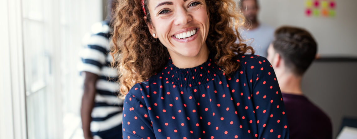 A cheerful woman with curly hair and a red headband smiles warmly at the camera, embodying confidence and self-compassion. She wears a navy blouse with red polka dots, standing with arms crossed in a bright office space. In the background, blurred colleagues and colorful notes create a supportive, collaborative environment, ideal for practicing self-compassion exercises.