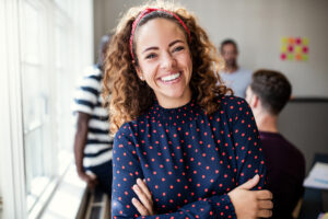 A cheerful woman with curly hair and a red headband smiles warmly at the camera, embodying confidence and self-compassion. She wears a navy blouse with red polka dots, standing with arms crossed in a bright office space. In the background, blurred colleagues and colorful notes create a supportive, collaborative environment, ideal for practicing self-compassion exercises.