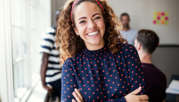 A cheerful woman with curly hair and a red headband smiles warmly at the camera, embodying confidence and self-compassion. She wears a navy blouse with red polka dots, standing with arms crossed in a bright office space. In the background, blurred colleagues and colorful notes create a supportive, collaborative environment, ideal for practicing self-compassion exercises.