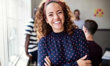 A cheerful woman with curly hair and a red headband smiles warmly at the camera, embodying confidence and self-compassion. She wears a navy blouse with red polka dots, standing with arms crossed in a bright office space. In the background, blurred colleagues and colorful notes create a supportive, collaborative environment, ideal for practicing self-compassion exercises.