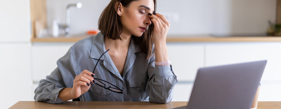 A young woman in a gray shirt sits at a desk with a laptop, rubbing the bridge of her nose while holding her glasses, looking fatigued. Signs of depression can include persistent exhaustion, trouble focusing, and feeling overwhelmed.