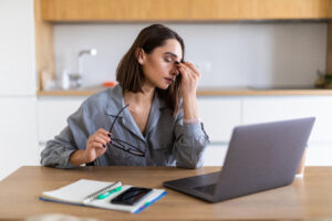 A young woman in a gray shirt sits at a desk with a laptop, rubbing the bridge of her nose while holding her glasses, looking fatigued. Signs of depression can include persistent exhaustion, trouble focusing, and feeling overwhelmed.