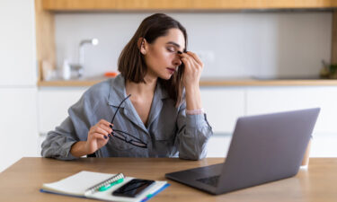 A young woman in a gray shirt sits at a desk with a laptop, rubbing the bridge of her nose while holding her glasses, looking fatigued. Signs of depression can include persistent exhaustion, trouble focusing, and feeling overwhelmed.