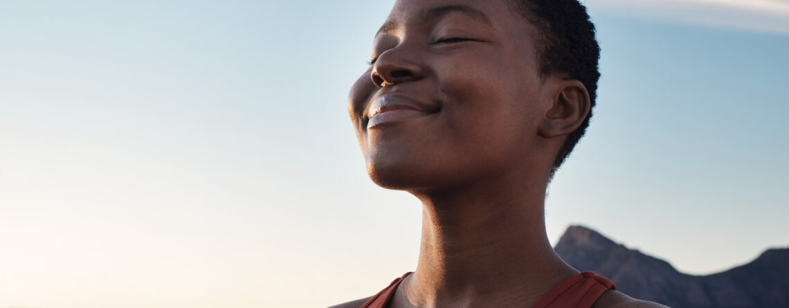 A Black woman in an orange tank top taking in a mindful breath, practicing mindfulness meditation with the sky as a background.