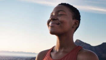 A Black woman in an orange tank top taking in a mindful breath, practicing mindfulness meditation with the sky as a background.