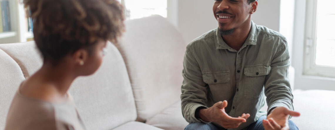 Cheerful positive handsome young african american guy in casual outfit sitting on couch at cozy counselor office, having conversation with therapist black woman, sharing good because he decided to start seeing a therapist