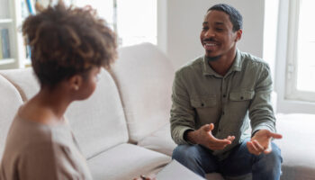 Cheerful positive handsome young african american guy in casual outfit sitting on couch at cozy counselor office, having conversation with therapist black woman, sharing good because he decided to start seeing a therapist