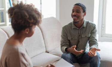 Cheerful positive handsome young african american guy in casual outfit sitting on couch at cozy counselor office, having conversation with therapist black woman, sharing good because he decided to start seeing a therapist