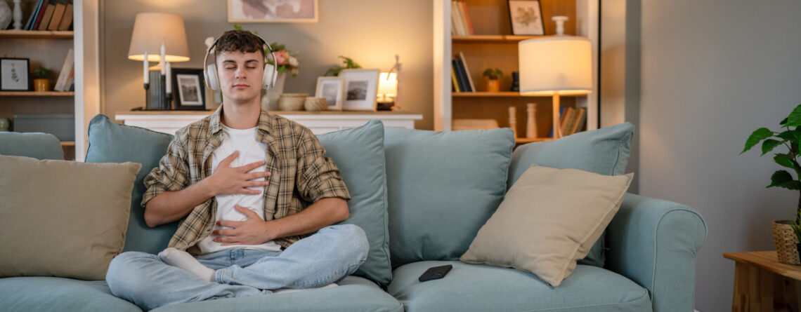 A young man wearing headphones sits cross-legged on a couch with his hands on his chest and stomach, practicing deep breathing as a strategy to manage anxiety.