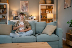 A young man wearing headphones sits cross-legged on a couch with his hands on his chest and stomach, practicing deep breathing as a strategy to manage anxiety.