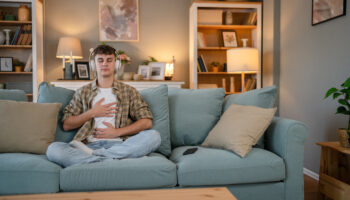 A young man wearing headphones sits cross-legged on a couch with his hands on his chest and stomach, practicing deep breathing as a strategy to manage anxiety.