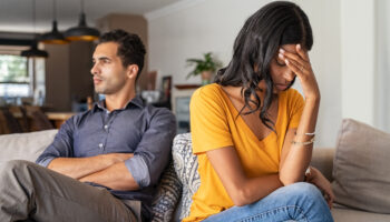 Young couple sitting on couch after an argument. Sad woman sitting with hand on head after quarrel with boyfriend at home. Angry couple ignoring each other on the sofa, having relationship troubles.