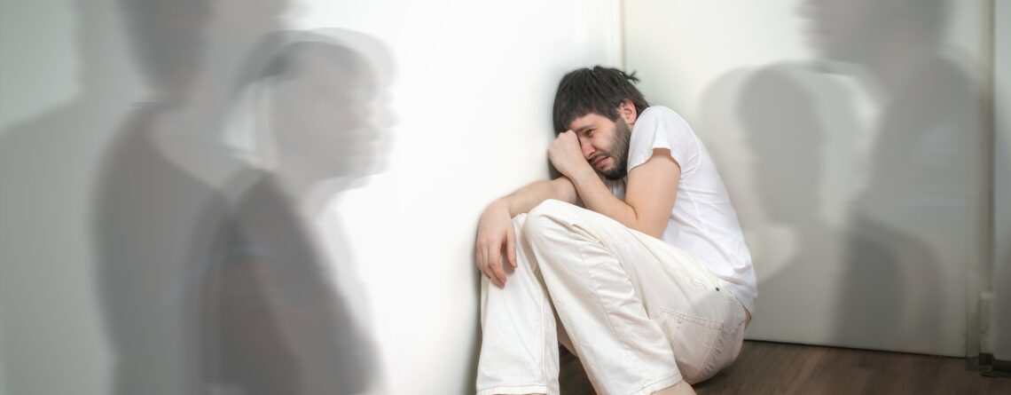 Scared and frightened man sitting in corner wearing white clothes has hallucinations and sees ghosts showing schizophrenia symptoms